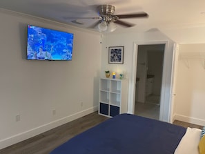 Ein Schlafzimmer mit einem Bett, einem Deckenventilator, einem an der Wand befestigten Fernseher, einer Tür und einem kleinen Regal mit Dekoration.