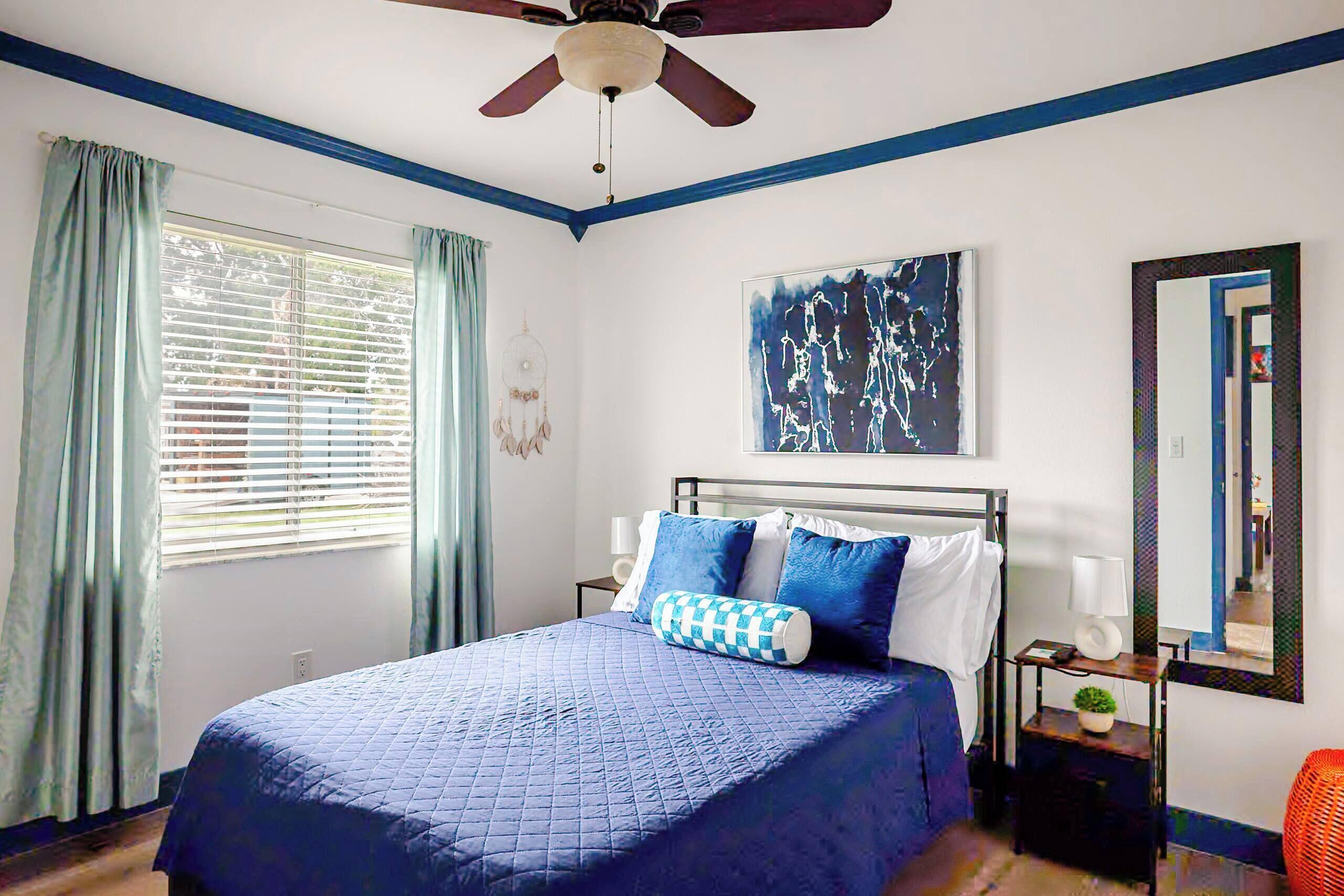 Une chambre à coucher comprenant un lit, un ventilateur de plafond, une fenêtre avec des rideaux, une œuvre d’art accrochée au mur et un miroir.
