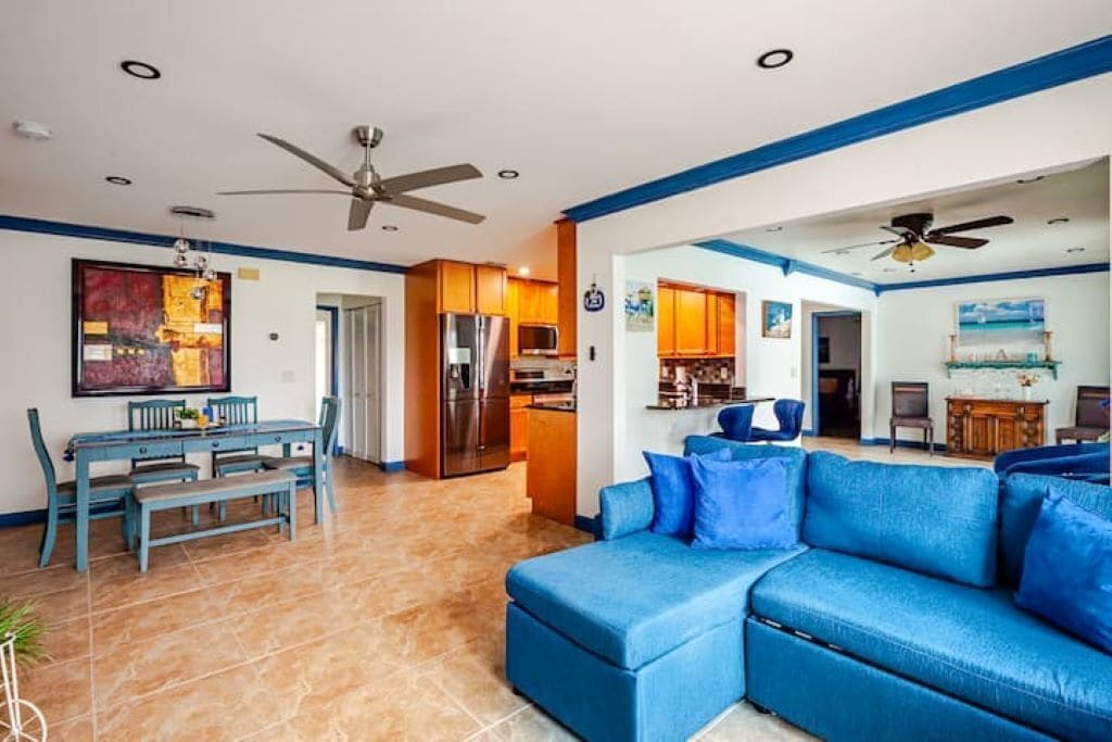 Uma sala de estar ampla com um sofá azul, uma mesa de jantar com cadeiras e uma cozinha ao fundo.