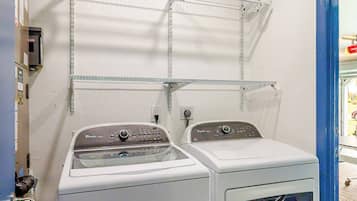 A laundry room with two white washing machines, metal shelves above, and a blue door to the right.