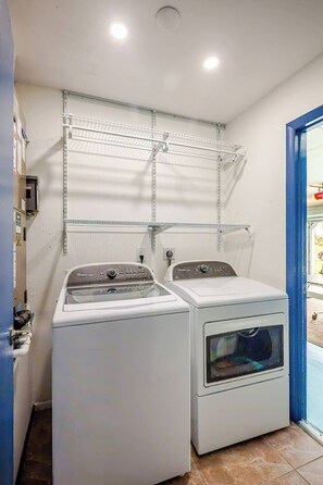 Une buanderie avec deux machines à laver blanches, des étagères métalliques au-dessus et une porte bleue sur le côté droit.