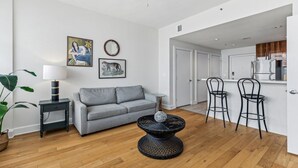 Ein modernes Wohnzimmer mit einem grauen Sofa, einem runden Couchtisch und einem Deckenventilator. Es gibt eine Küchenzeile mit einer weißen Arbeitsplatte und Barhocker.
