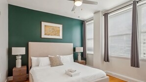 Un dormitorio ordenado con una cama grande, mesitas de noche con lámparas, un ventilador de techo y una ventana con persianas.