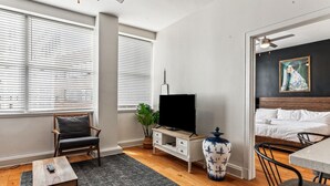 Un salon moderne avec un téléviseur à écran plat, une table basse en bois, un fauteuil, un petit meuble blanc, un vase décoratif et un lit avec une tête de lit sombre.