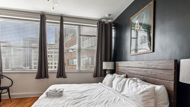 Une chambre d’hôtel moderne avec un grand lit, une tête de lit en bois, une vue sur la ville grâce à de grandes fenêtres du sol au plafond et un tableau accroché au mur.