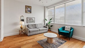 Un salon avec un canapé gris, un fauteuil turquoise, une table basse ronde, un ventilateur de plafond et une grande fenêtre avec des stores.