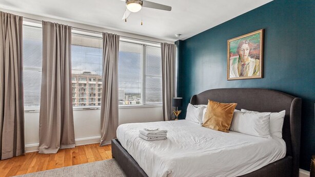 Une chambre à coucher avec un lit, des rideaux, un ventilateur de plafond, un tableau et une vue sur les bâtiments par la fenêtre.