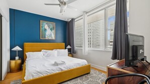 Un dormitorio con una cama, ventilador de techo, escritorio con computadora y ventana con persianas.