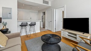 Una sala moderna con sofá, televisor, zona de comedor con sillas y una cocina al fondo.