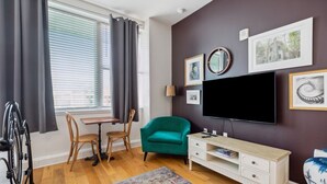 A room with a large window, a teal armchair, a white TV stand, and a wheelchair. - Urban Retreat Escape Studio (New Orleans)