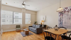 Un salon moderne avec un canapé, une table à manger et des chaises. On y trouve un ventilateur de plafond, un tableau au mur et une fenêtre avec des stores.