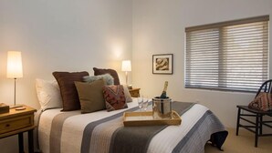 Un dormitorio con cama, mesitas de noche, lámparas, una ventana con persianas y una silla.