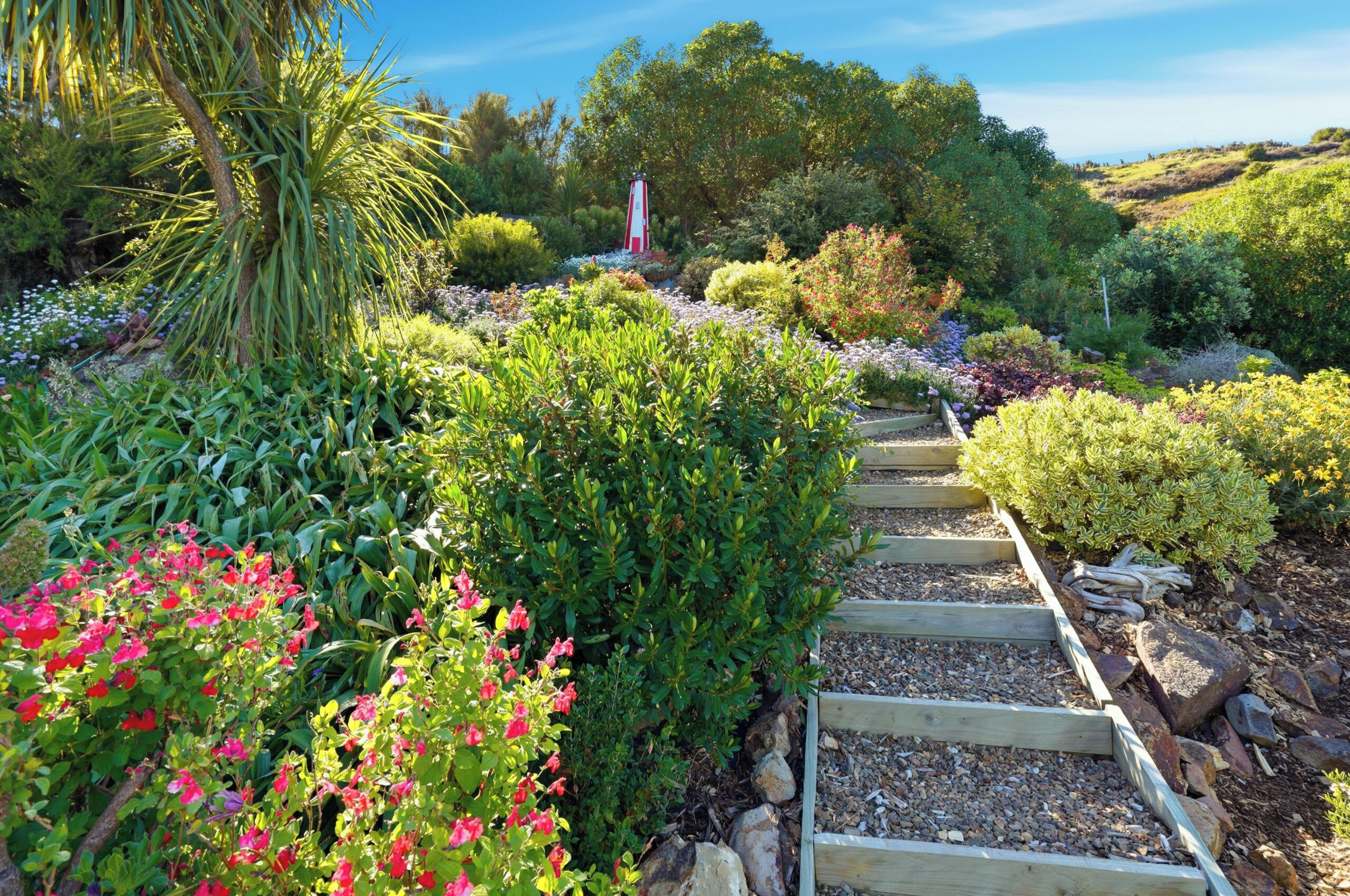 A garden pathway with various plants and a lighthouse in the background.