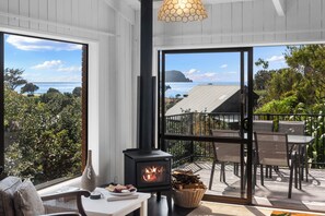 A cozy room with a wood-burning stove, a dining area with chairs and a table, and a large window offering a view of the sea and greenery. - Seaview Family Haven - Pauanui Holiday Home (Pauanui)