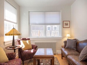 A living room with a sofa, armchair, and a small table. There is a window with blinds, a radiator, and a lamp on a side table.