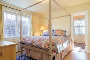 Een bed met hemelbed met bloemenbeddengoed, een houten nachtkastje en een raam met gordijnen.