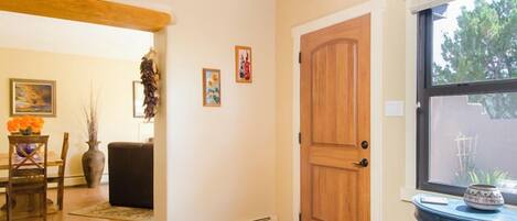 A room with a wooden door, a window with a view, a table with a vase, and a chair.
