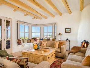 A cozy living room with a fireplace, a sofa set, a wooden coffee table, and large windows offering a view.