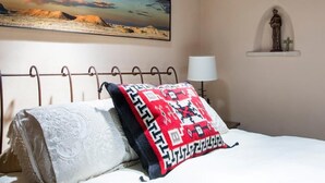 Una cama con un cojín estampado y una cubierta de encaje, una mesita de noche con una lámpara, un estante en la pared con una estatua y un cuadro de un paisaje desértico.
