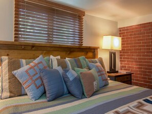 A bedroom with a bed, pillows, a nightstand, a lamp, and a brick wall.