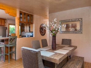 A dining area with a wooden table, cushioned chairs, a vase with flowers, and a framed picture on the wall.