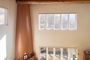 A room with a large chimney, a window with a view of trees, and a window with a view of a building.