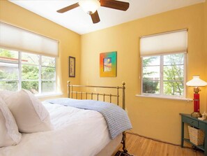 Una camera da letto con un letto, due finestre con persiane, una ventola a soffitto e un comodino con una lampada.