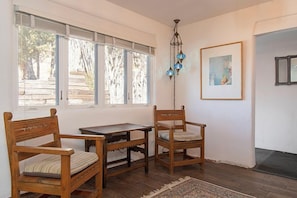A room with a wooden bench, a small table, and a chair. There is a window with blinds, a framed picture on the wall, and a door with a mat.