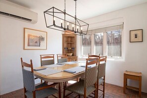 Zona pranzo con tavolo rotondo, sei sedie, lampadario e finestra con tende.