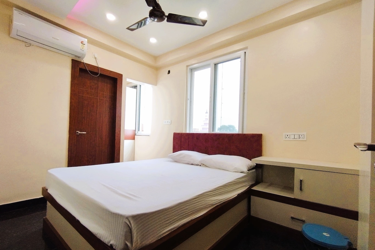 A hotel room with a bed, a ceiling fan, an air conditioner, a window, and a small desk with a trash bin.
