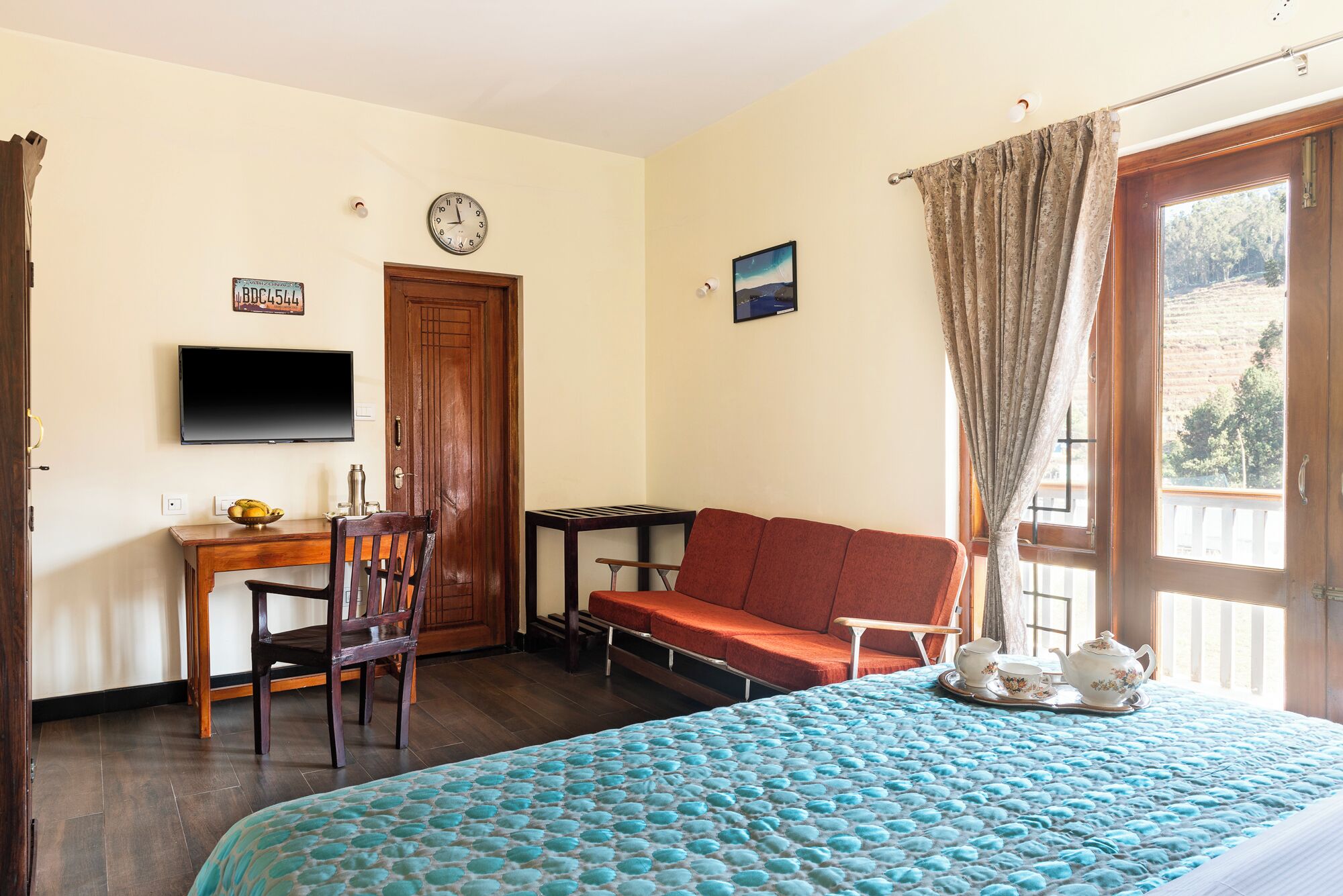 A room with a bed, a sofa, a TV, a desk with a chair, a clock, and a window with curtains.