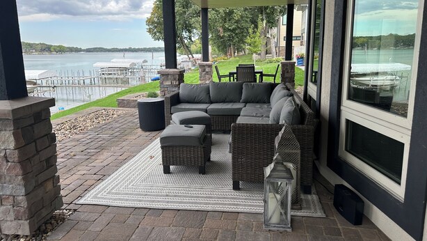 A covered patio with a sectional sofa, outdoor table, and chairs, overlooking a lake and dock area.