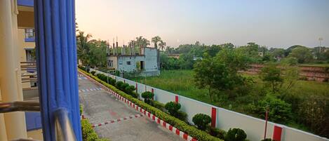 A view from a balcony overlooking a road with a red and white striped barrier, green bushes, and a building in the distance.