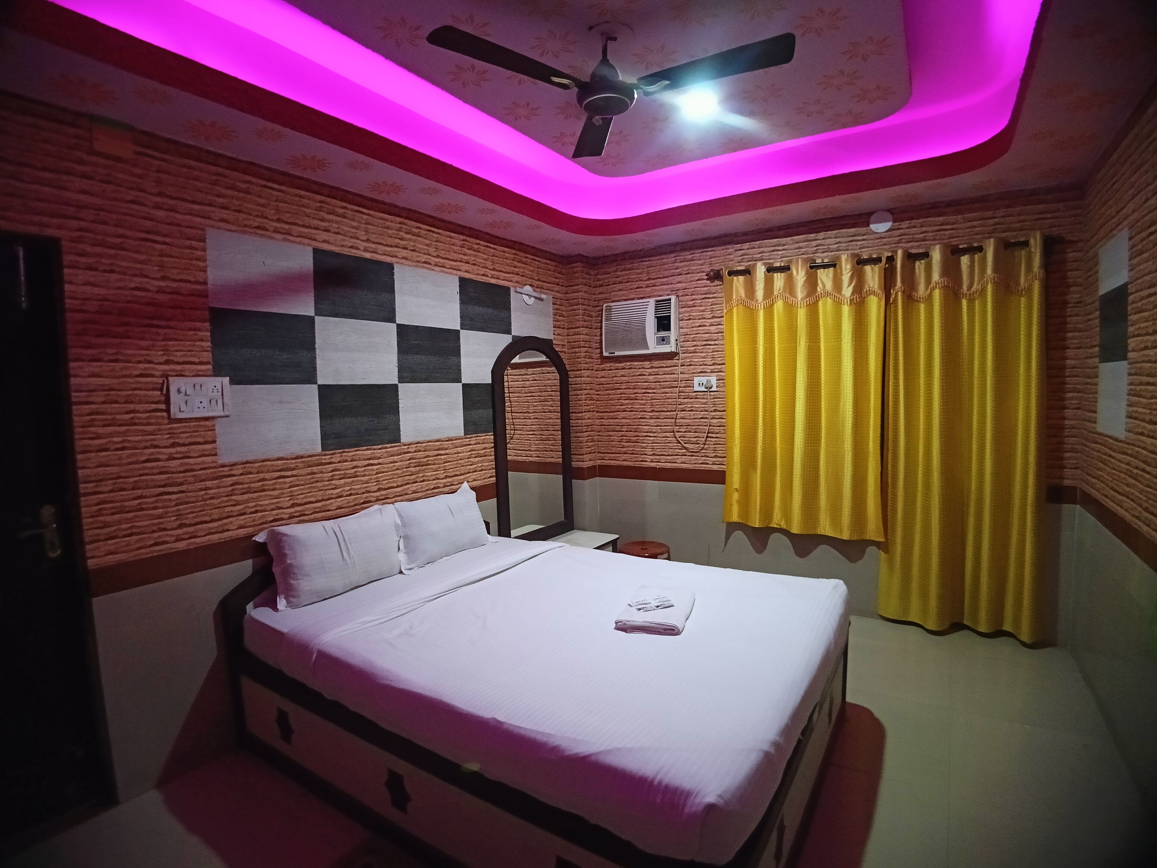 A bedroom with a bed, a ceiling fan, a mirror, and a window with yellow curtains.