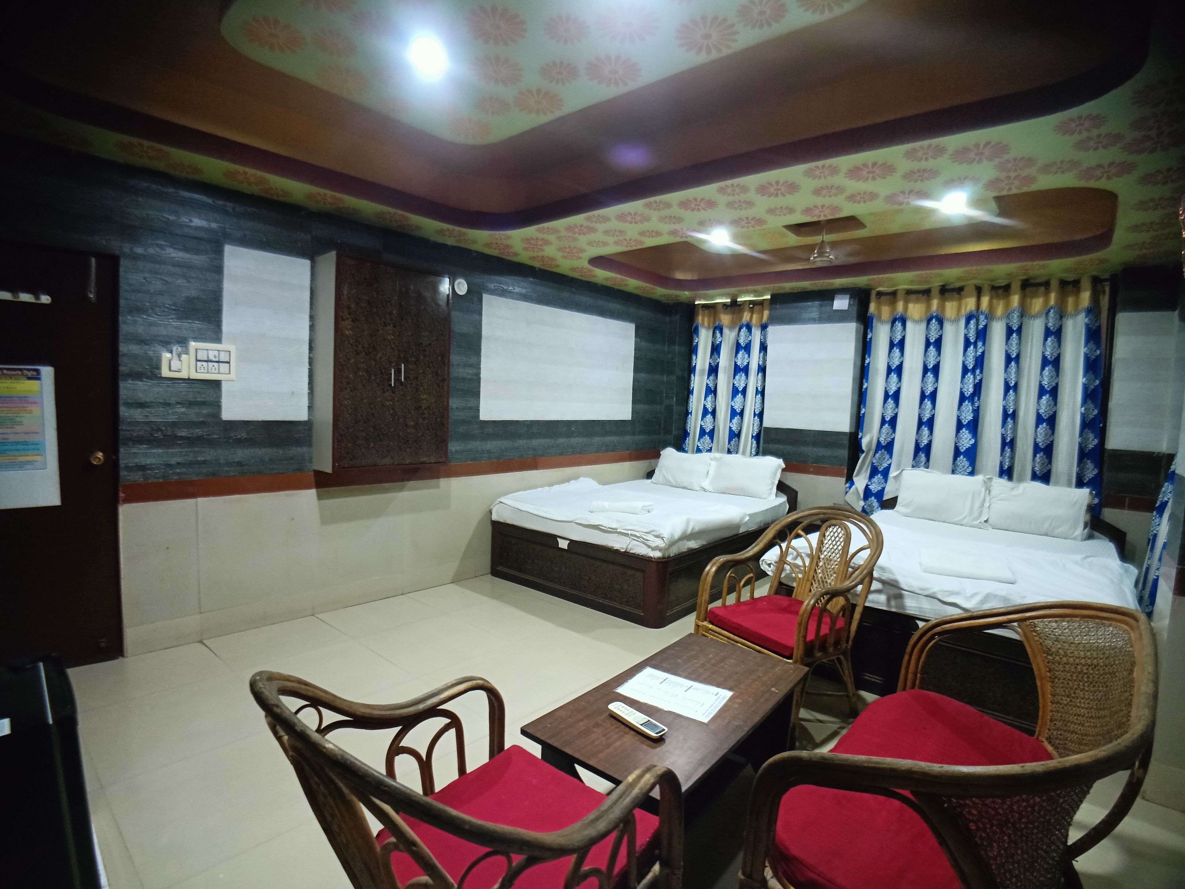 A hotel room with two single beds, a wooden table, and two wicker chairs with red cushions.