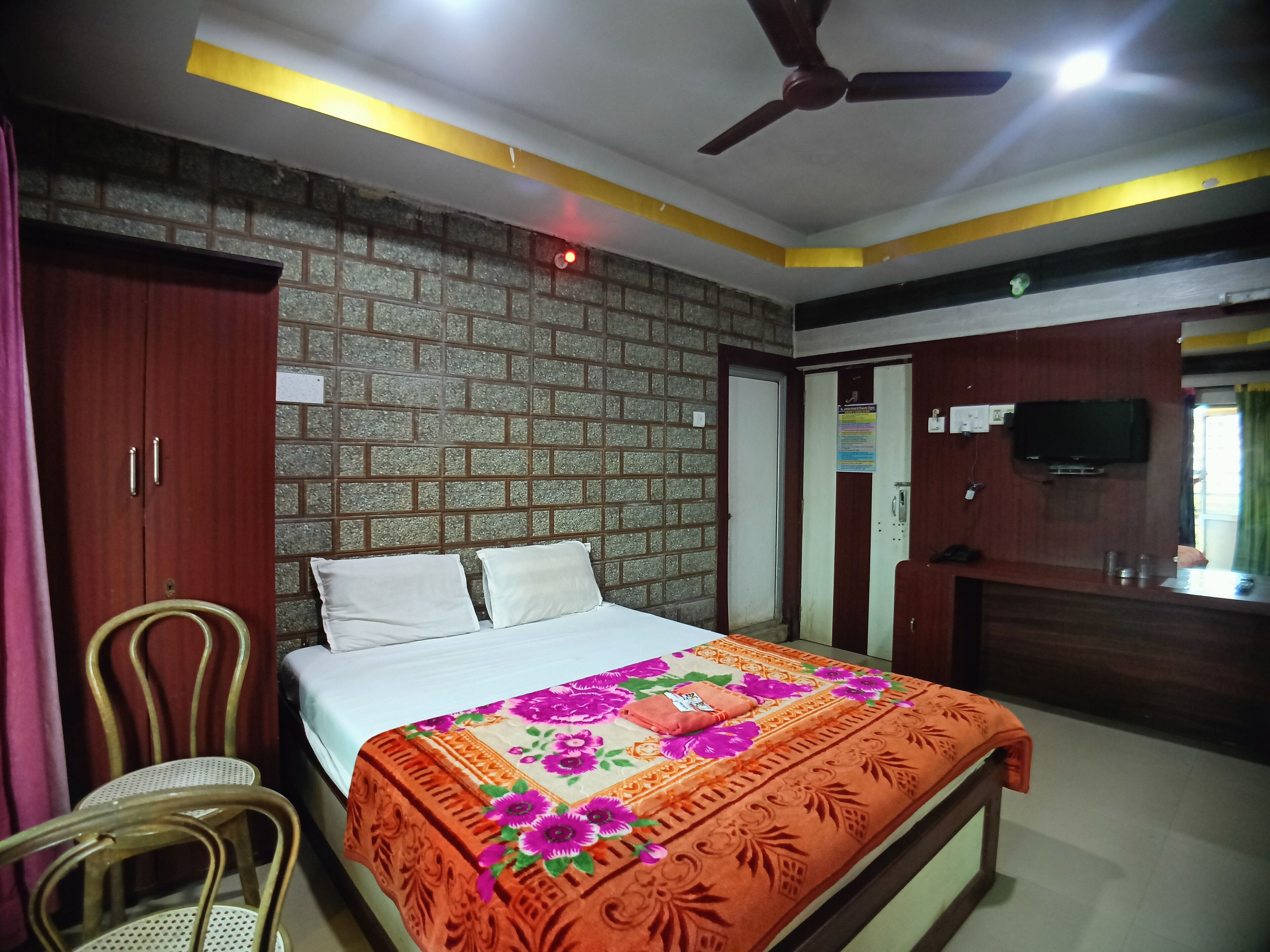 Une chambre d’hôtel avec un mur décoré d’un motif en briques, un lit recouvert d’une couvre-lit à motifs floraux, un ventilateur de plafond, un téléviseur fixé au mur et une armoire en bois.
