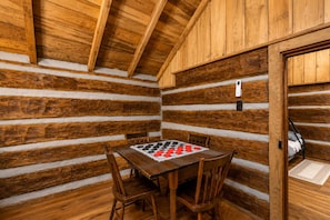 Interior de una cabaña de troncos con una mesa y sillas de madera, un tablero de juego con cuadros y una cama con cabecera blanca.