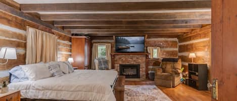 A log cabin bedroom with a bed, fireplace, TV, and a view of the outdoors.