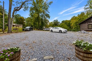 A gravel driveway with a white car parked on the right, a black truck on the left, and a wooden cabin in the background.