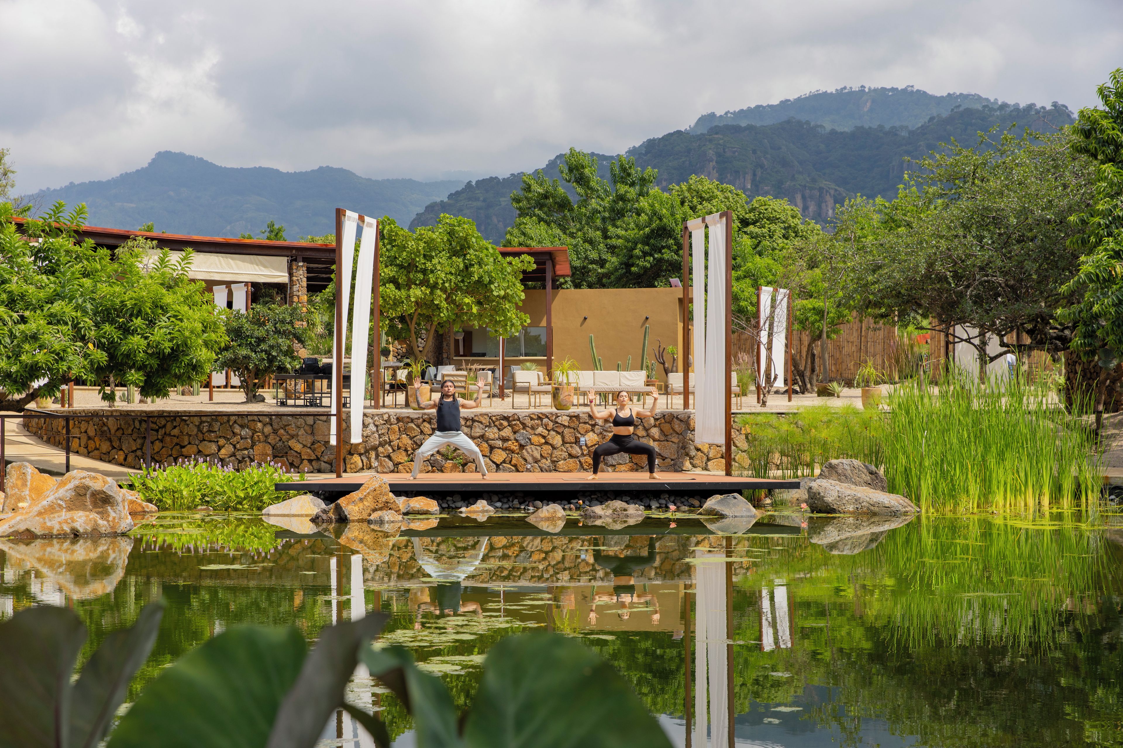 A tranquil outdoor yoga area with a pond, surrounded by a stone wall and lush greenery, overlooking a mountainous landscape.