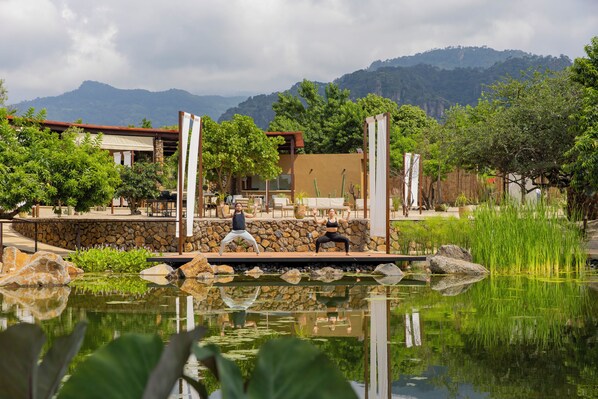 A tranquil outdoor yoga area with a pond, surrounded by a stone wall and lush greenery, overlooking a mountainous landscape.