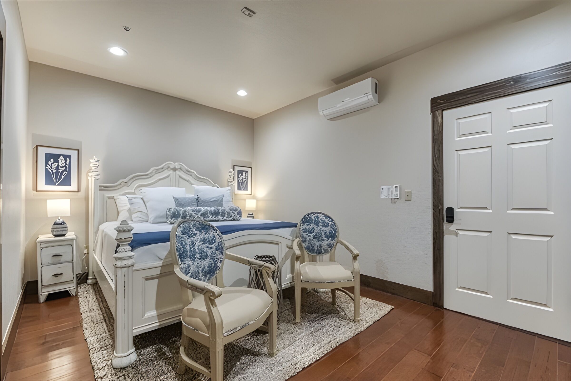 A bedroom with a bed, bedside tables, chairs, a rug, a door, and an air conditioning unit.