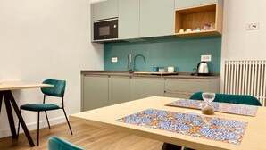 Een compacte keuken met blauwgroene kastjes, een magnetron en een spoelbak. Er staat een houten tafel met een kleurrijke placemat en een klein zandloper. Rond de tafel staan twee blauwgroene stoelen.