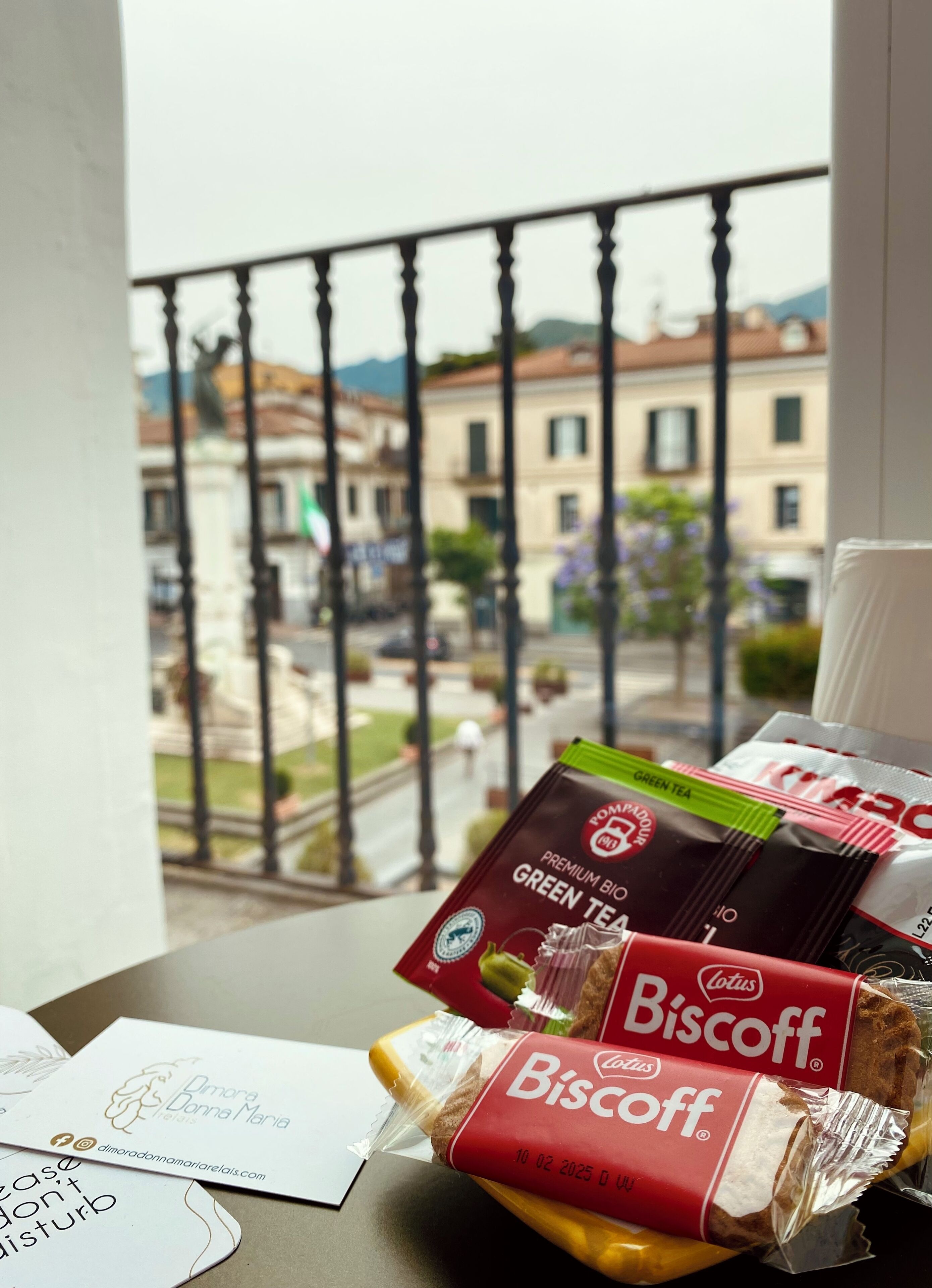 Un tavolo con una confezione di biscotti Biscoff, una bustina di tè verde e un biglietto da visita su un balcone con vista sulla città.