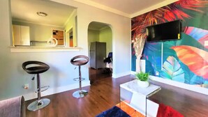 Una sala moderna con piso de madera, un cuadro abstracto colorido, un televisor montado en la pared y dos taburetes de bar.