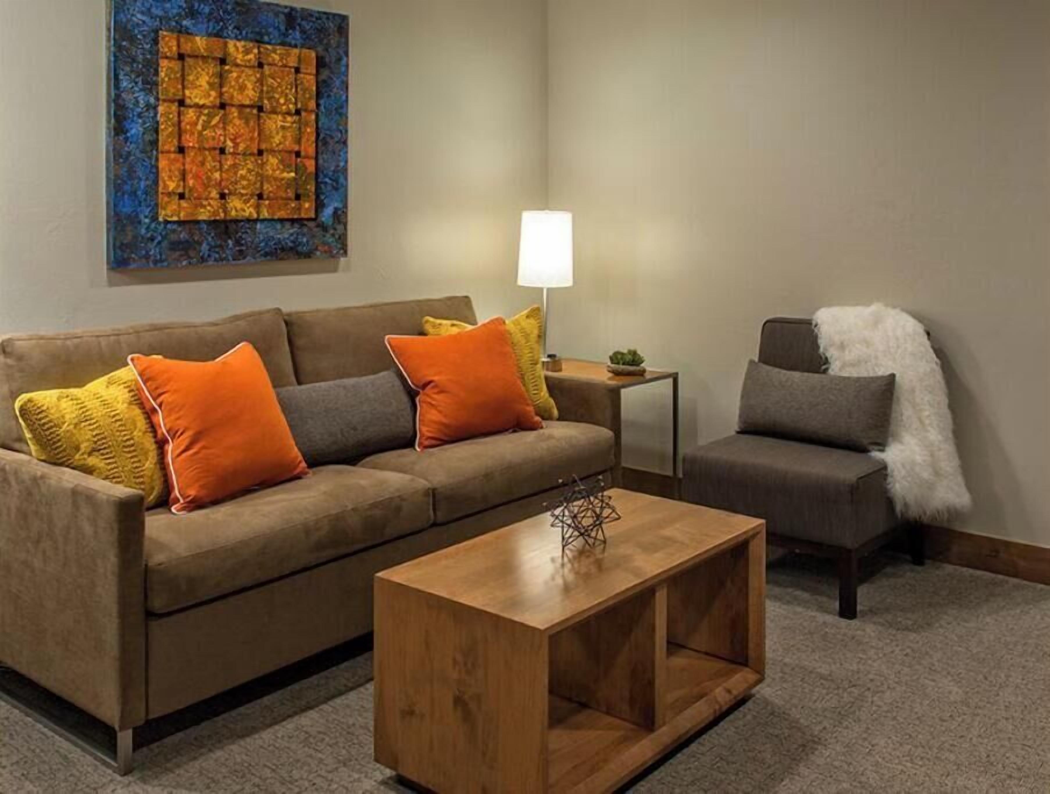 A modern living room with a brown sofa, a wooden coffee table, and a gray armchair. A blue abstract painting is on the wall, and a small table with a lamp is visible.