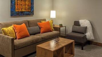 A modern living room with a brown sofa, a wooden coffee table, and a gray armchair. A blue abstract painting is on the wall, and a small table with a lamp is visible.