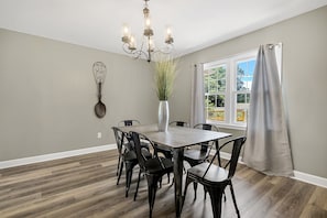 A dining room with a wooden table, black chairs, a chandelier, a vase with greenery, and a window with curtains. - American Retreat (Huntsville)