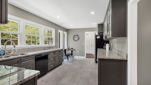 A kitchen with dark cabinets, a granite countertop, a dining area with two chairs, and a view through the window. - American Retreat (Huntsville)