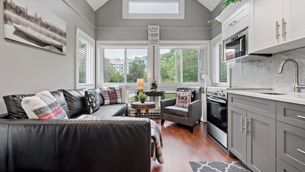Een moderne woonkamer met een bank, fauteuils en een eettafel. Er is een keuken met kasten en apparatuur. Een groot raam zorgt voor natuurlijk licht en uitzicht.